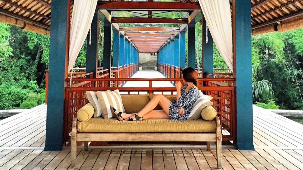 On a large canopied deck, Brooke is lounging on a couch in the foreground. The length of the deck tunnels into the distance, surrounded by forest trees. 